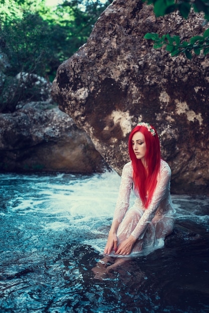 Belle jeune fille au repos dans l'eau. Jeune femme à la robe blanche est assise sur la pierre au milieu d'un ruisseau.