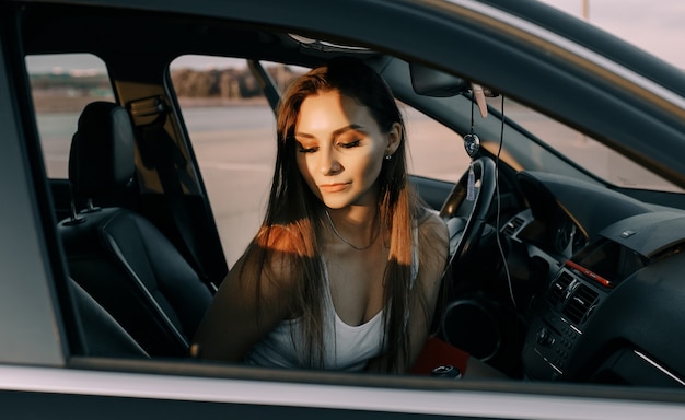 Belle jeune fille au volant d'une voiture le soir au coucher du soleil sur un parking vide