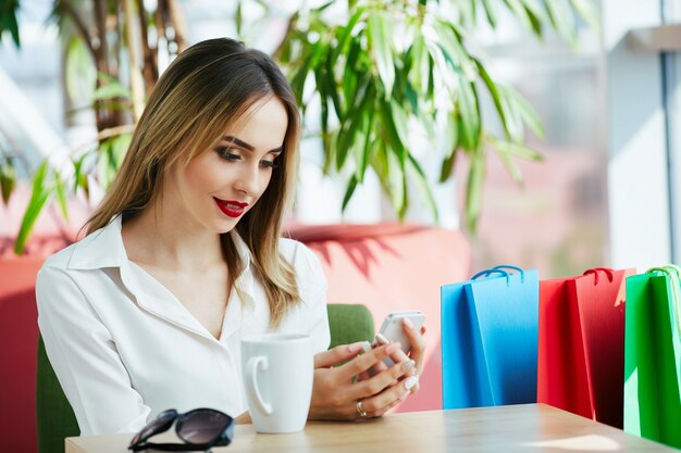 Belle jeune fille aux cheveux brun clair et aux lèvres rouges portant un chemisier blanc et assis avec des sacs colorés et une tasse de café, tenant un téléphone mobile.