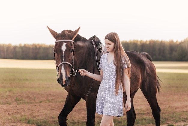 Une belle jeune fille aux cheveux longs marche à côté d'un cheval brun en été dans la nature.