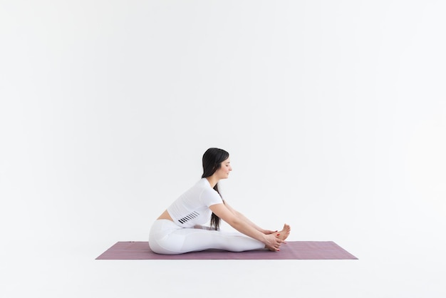 une belle jeune fille aux cheveux noirs se tient dans la pose Paschimottanasana sur fond blanc