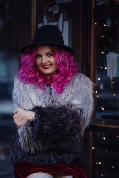 Photo une belle jeune fille aux cheveux roses et un manteau inhabituel se promène dans la ville du soir profite de la lumière des guirlandes et de la lumière du coucher du soleil