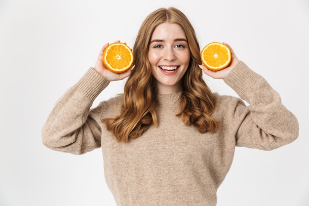 Belle jeune fille aux longs cheveux blonds bouclés portant un pull debout isolé sur un mur blanc, montrant des tranches d'orange