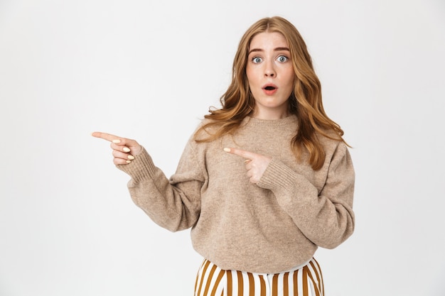 Belle jeune fille aux longs cheveux bouclés blonds portant un pull debout isolé sur un mur blanc, pointant vers l'espace de copie