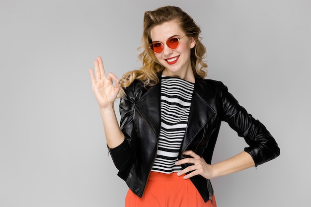 Belle jeune fille blonde en blouse rayée et veste en cuir souriant dans des lunettes de soleil avec les mains sur la taille debout