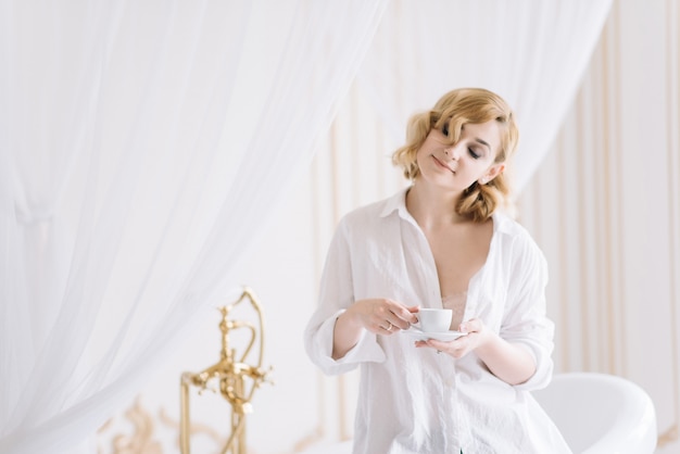 Belle jeune fille blonde dans une chemise légère dans une pièce lumineuse bénéficie d'une tasse de café le matin avant d'aller travailler.Fille dans la salle de bain dans des vêtements candides.