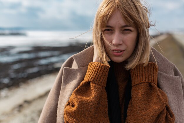 Une belle jeune fille blonde dans un pull marron et un manteau beige se dresse sur une route vide