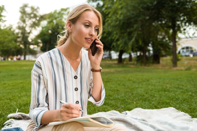 Belle jeune fille blonde se reposant sur une pelouse dans le parc, prenant des notes dans un journal tout en parlant au téléphone portable