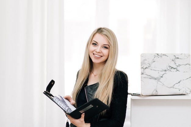 Belle Jeune Fille Blonde Souriante Avec Un Ordinateur Portable Et Un Stylo Dans Ses Mains, à Côté D'un Ordinateur Portable Sur Une Fenêtre Lumineuse, Employé De Bureau