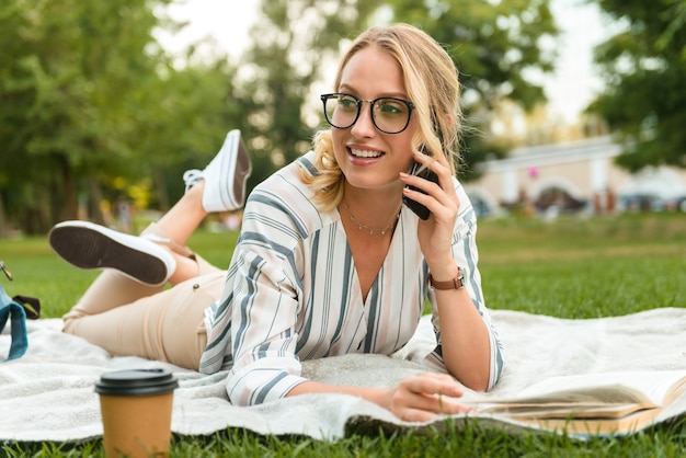 Belle jeune fille blonde souriante se reposant sur une pelouse dans le parc, parlant au téléphone portable tout en s'étendant sur une couverture