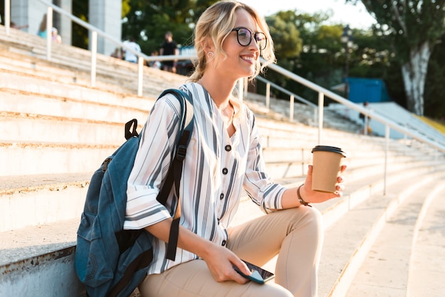 Belle jeune fille blonde utilisant un téléphone portable tout en étant assise sur des marches à l'extérieur et en buvant du café