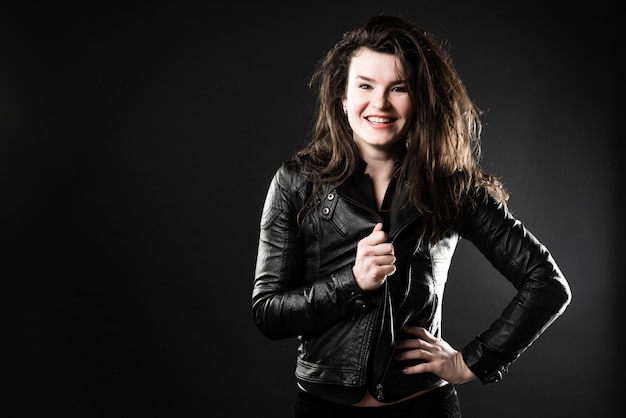 Belle jeune fille brune dans une veste noire pose en studio sur un fond sombre.