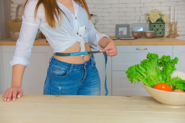 Belle jeune fille brune sportive avec un torse nu mesurant sa taille avec un ruban à mesurer dans la cuisine. Légumes frais sur la table. Femme fitness