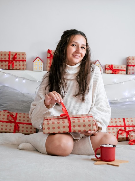 Belle jeune fille brune vêtue d'une robe en tricot blanc et de chaussettes en laine est assise sur le lit avec une boîte-cadeau La chambre est décorée de guirlandes et de boîtes-cadeaux Atmosphère de maison confortable