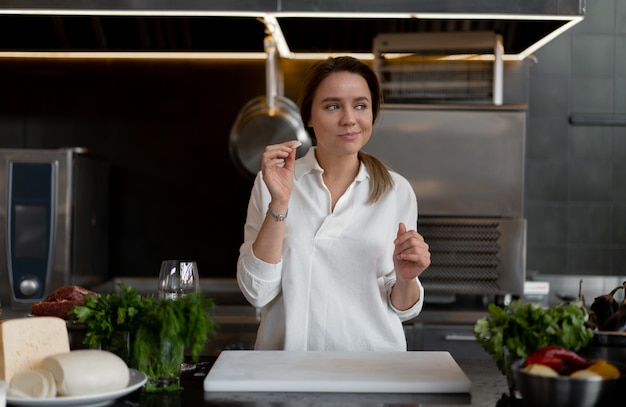 Belle jeune fille caucasienne debout dans la cuisine dans un uniforme blanc souriant et dégustant un morceau de fromage. Jolie femme des années 30 en chemise blanche avec de la nourriture Ingrédients fromage légumes viande.