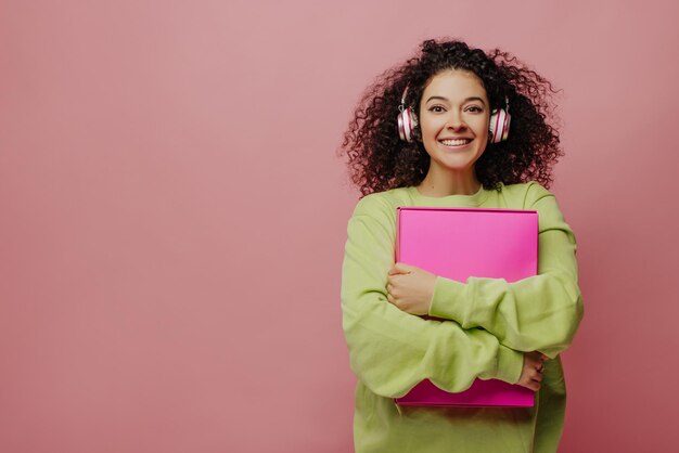 Belle jeune fille caucasienne porte des écouteurs et tient la boîte sur fond rose avec un espace pour le texte Brunette aux cheveux bouclés en chandail vert Concept de musique de technologie audio