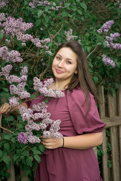 Une belle jeune fille caucasienne souriante heureuse en robe violette près d'un buisson de lilas en fleurs. Modèle plus taille. Printemps, style rustique.