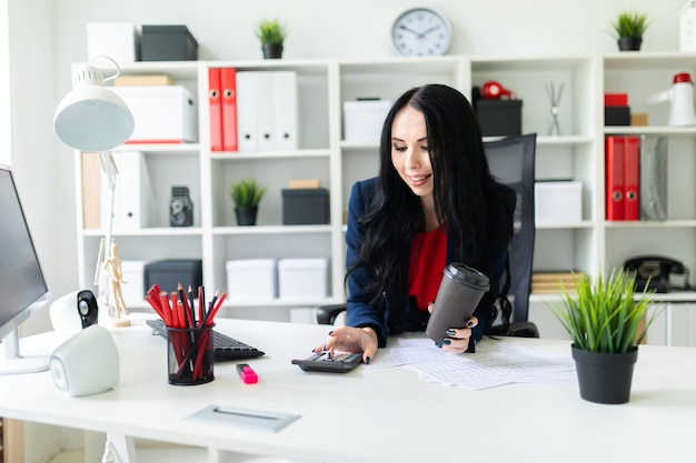 Une belle jeune fille compte avec la main sur la calculatrice, tient dans sa seconde main un verre à café au bureau à la table.