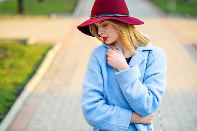 Belle jeune fille dans un manteau bleu et un chapeau bordeaux marchant dans la rue par une journée de printemps ensoleillée