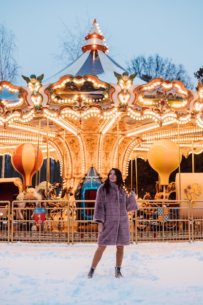 Une belle jeune fille dans un manteau de fourrure se tient près du carrousel un soir d'hiver