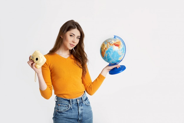 Une belle jeune fille dans un pull jaune tient un globe terrestre et un appareil photo. Loisirs, voyages, tourisme. Espace pour le texte