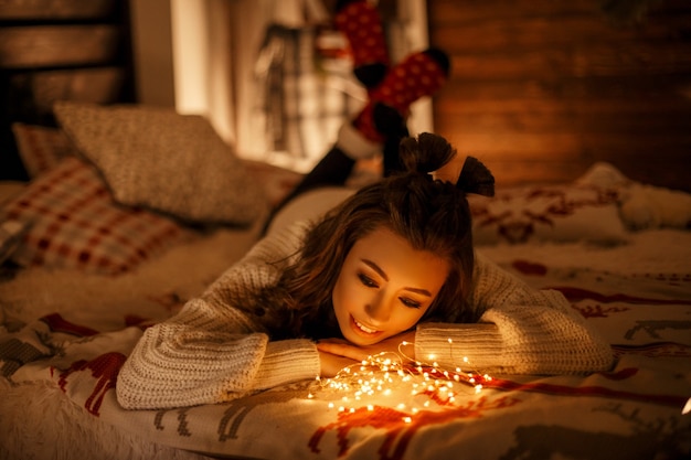 Belle jeune fille dans un pull vintage tricoté avec des lumières festives sur un lit la veille de Noël