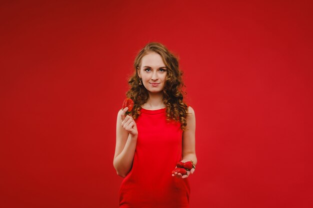 Une belle jeune fille dans une robe rouge sur un rouge tient une fraise dans ses mains et sourit.