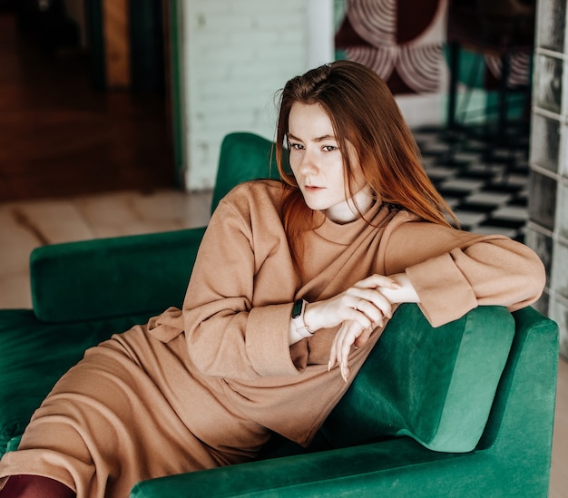 Belle jeune fille élégante aux cheveux roux est assise sur un canapé vert à l'intérieur dans des vêtements chauds dans une atmosphère chaleureuse avec un intérieur élégant