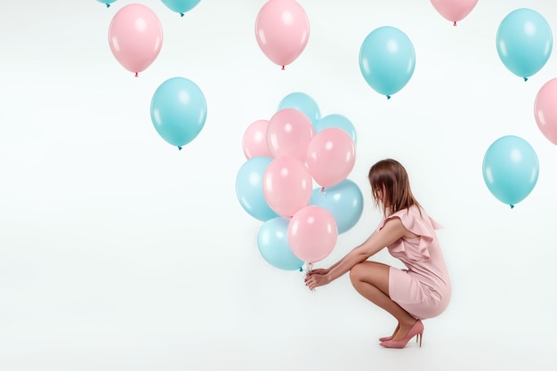 Une belle jeune fille est assise avec des ballons bleus et roses sur un mur blanc.