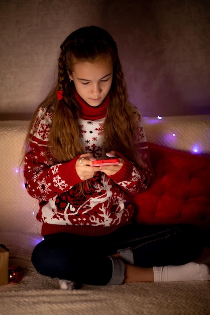 Une belle jeune fille est assise sur le canapé et regarde le téléphone portable Noël Nouvel An