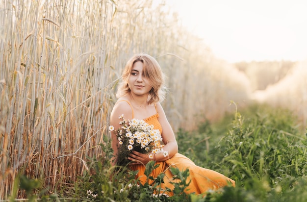 Une belle jeune fille est assise sur un champ de blé et tient un bouquet de marguerites dans ses mains.