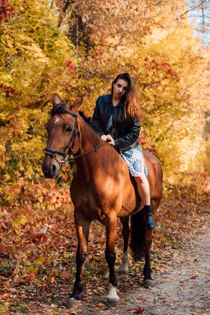 Une belle jeune fille est assise à cheval sur un cheval vêtu d'une robe et de bottes et a l'air droite