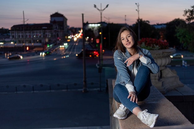 Belle jeune fille est assise seule sur une haute marche. Une étudiante souriante se repose. Ville de nuit en arrière-plan.