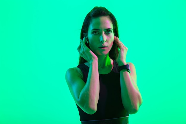 Belle jeune fille fitness debout isolée sur un mur de néon vert, portant des écouteurs sans fil