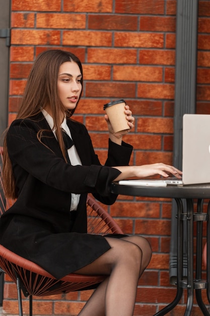 Belle jeune fille gestionnaire s'assoit et travaille sur son ordinateur portable en buvant du café près d'un mur de briques dans un café Femme d'affaires prospère se détendre et travailler dans la ville