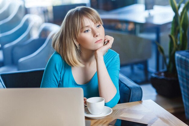 Une belle jeune fille indépendante réfléchie aux cheveux blonds coupés en chemisier bleu est assise dans un café et rêve, a une nouvelle idée et planifie sa propre stratégie, tenant une main sur le menton.