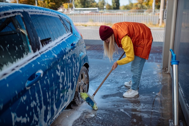 Belle jeune fille lave la voiture au lave-auto en libre-service en effaçant la saleté de la voiture avec une brosse