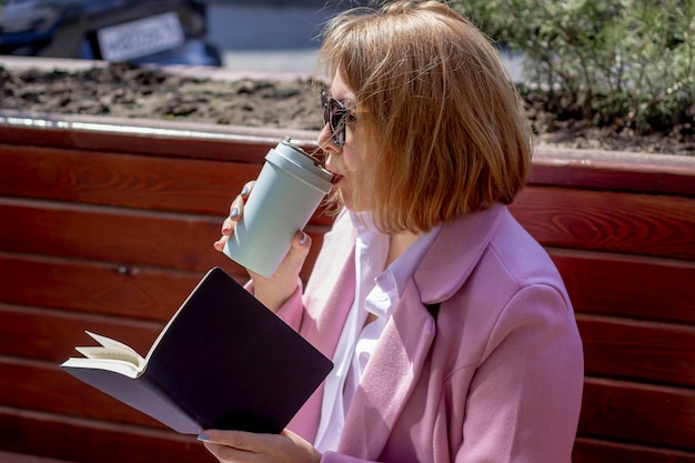 Une belle jeune fille à lunettes de soleil boit du café dans une thermotasse écologique et tient un cahier tout en étant assise sur un banc à l'extérieur par temps chaud