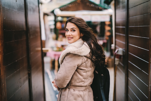 Belle Jeune Fille En Manteau D'hiver Debout Dans Le Passage De La Rue. Regardant La Caméra. Belle Rue Décorée De Noël.