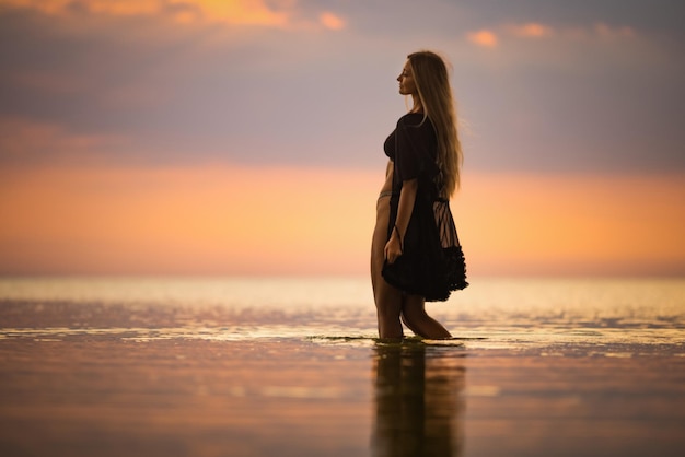 Une belle jeune fille mince aux cheveux blonds chics dans un maillot de bain bikini noir et un châle en dentelle noire, marche le long d'un estuaire peu profond sur fond d'un chaud coucher de soleil d'été ensoleillé