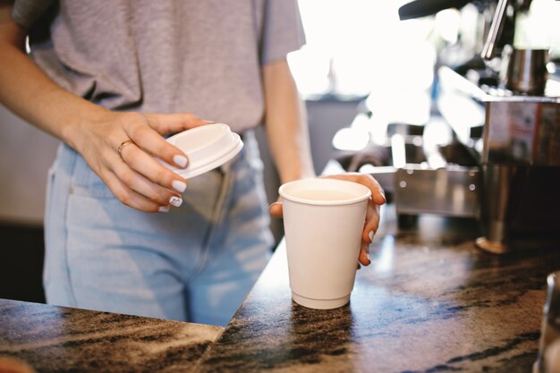 Une belle jeune fille mince, vêtue d'une tenue décontractée, met doucement le couvercle sur le café dans un café confortable. .