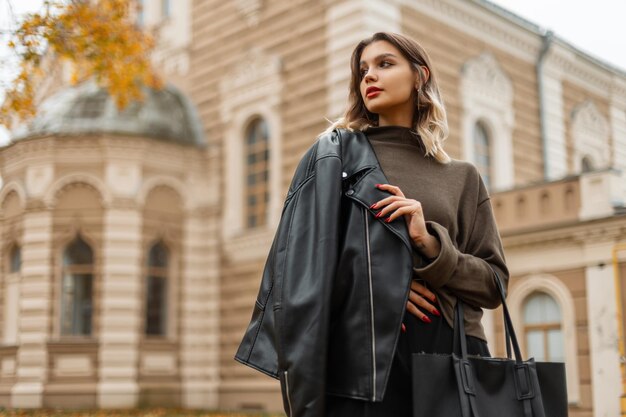 Belle jeune fille à la mode dans une élégante veste en cuir noir avec un pull vert avec un sac se promène dans la ville d'automne avec un bâtiment vintage