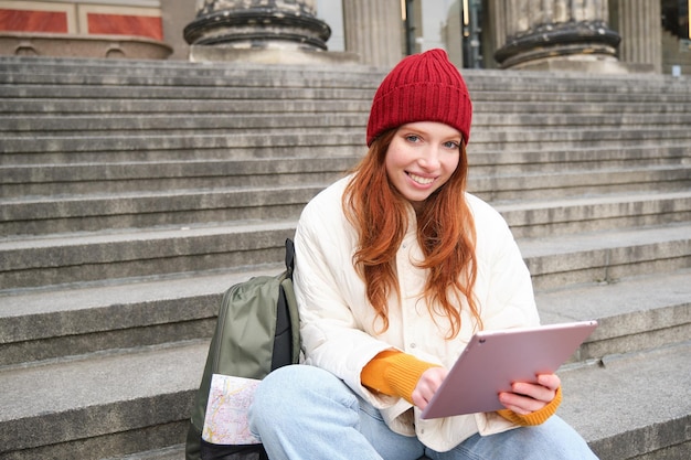 Belle jeune fille moderne aux cheveux rouges détient une tablette numérique se trouve dans les escaliers près du musée et connec