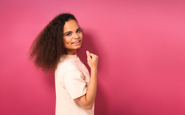 Belle jeune fille à moitié tourné à la recherche positive à l'avant portant un t-shirt pêche montrant ses muscles isolés sur un mur rose