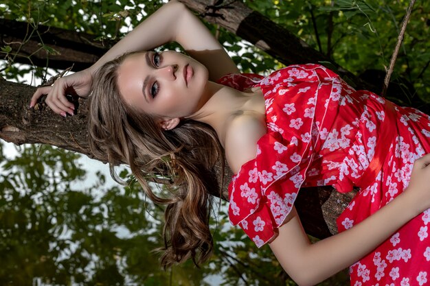 Une belle jeune fille à la peau parfaite, aux cheveux longs et à la robe rouge, repose sur une branche d’arbre à l’air détendu et languissant.