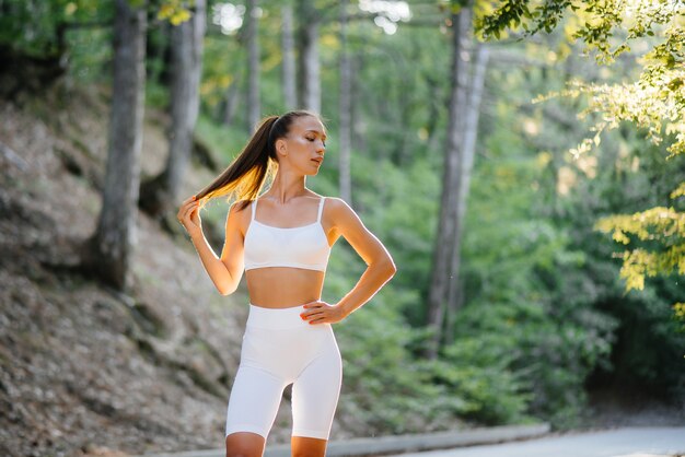 Une belle jeune fille pose avant de s'entraîner, sur la route dans une forêt dense, au coucher du soleil. Un mode de vie sain et courir au grand air.