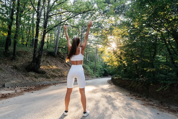 Une belle jeune fille pose avec son dos avant de s'entraîner sur la route dans une forêt dense au coucher du soleil Un mode de vie sain et courir à l'air frais