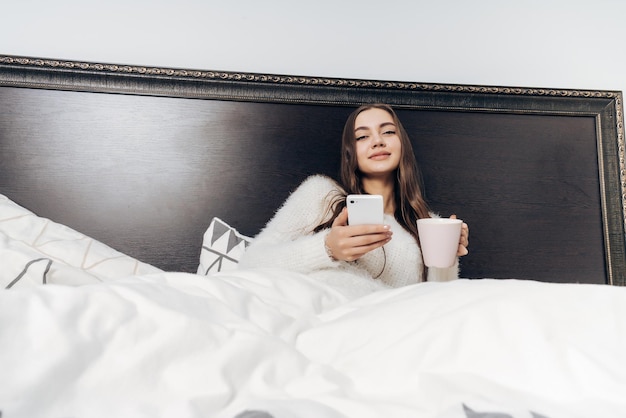 Belle jeune fille en pyjama couché dans son lit le soir se reposer après une dure journée de travail et tenant un smartphone