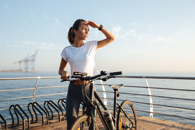 Belle jeune fille de remise en forme debout à l'extérieur avec un vélo, paysage de mer