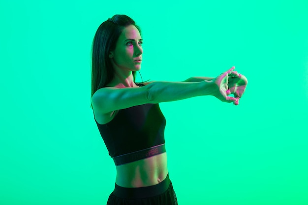 Belle jeune fille de remise en forme debout isolée sur un mur de néon vert, faisant des exercices d'étirement